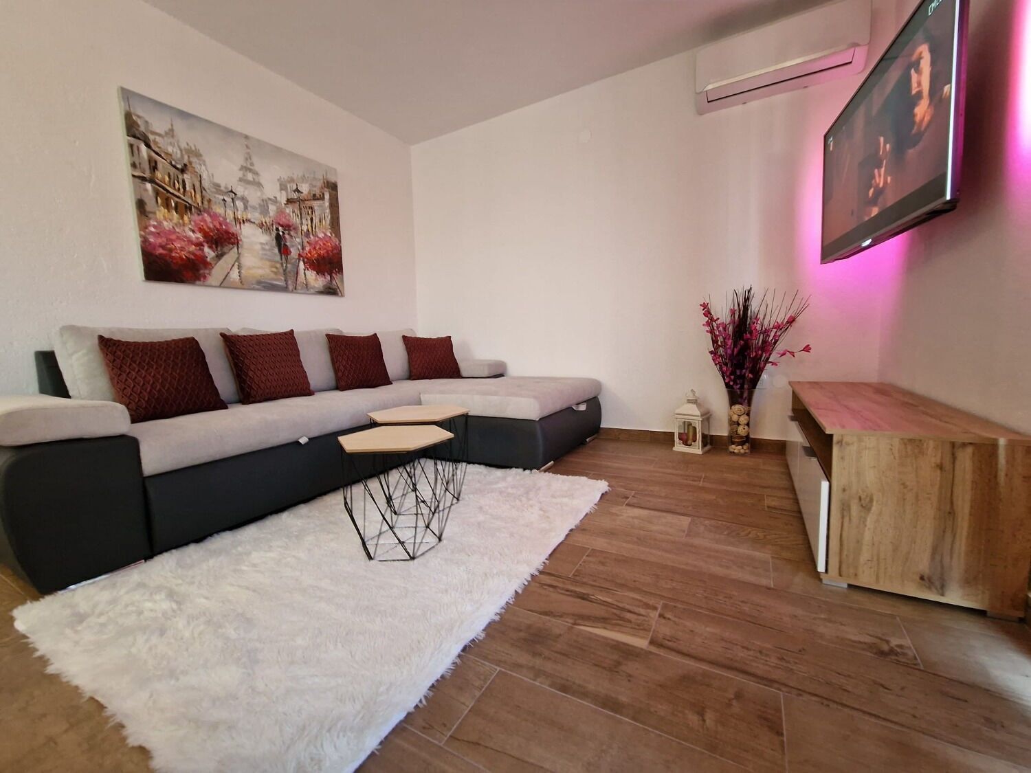 Una sala moderna con un sofá seccional, una alfombra mullida blanca, una mesa de centro de madera, una mesita auxiliar y un cuadro en la pared.