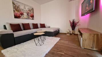 Una sala moderna con un sofá seccional, una alfombra mullida blanca, una mesa de centro de madera, una mesita auxiliar y un cuadro en la pared.