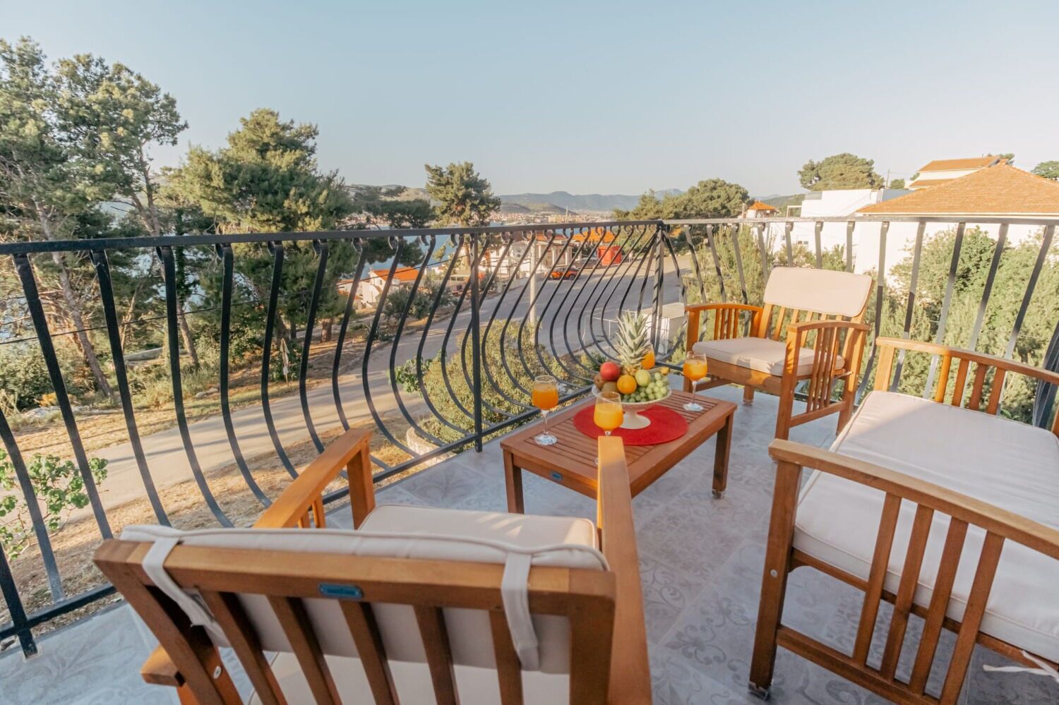 Een balkon met houten meubels, een tafel met fruit en drankjes, en uitzicht op een stadsbeeld.