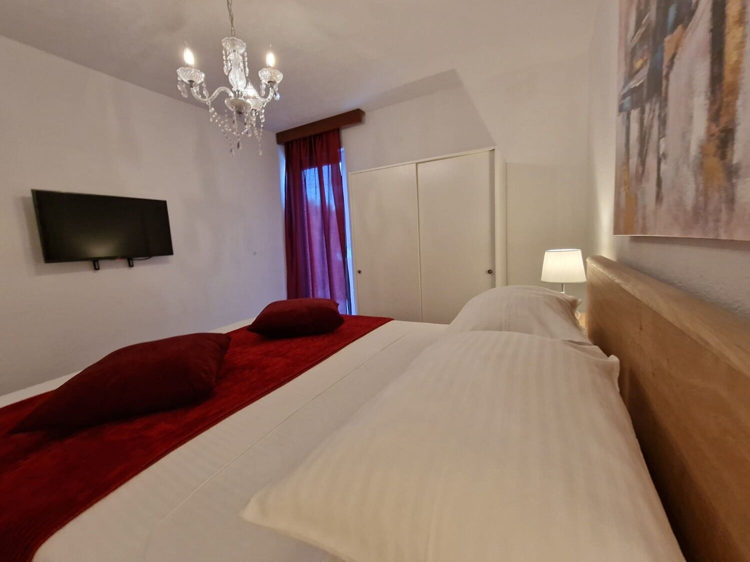 A hotel room with a bed, a TV, a chandelier, a painting, and a door.