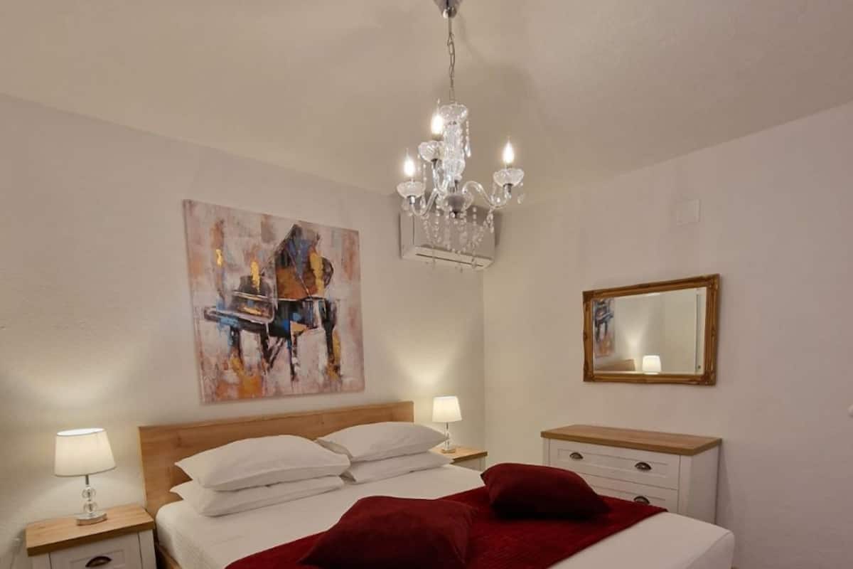 Un dormitorio con cama de madera, ropa de cama blanca, manta roja, candelabro, una pintura, espejo y suelo de madera.