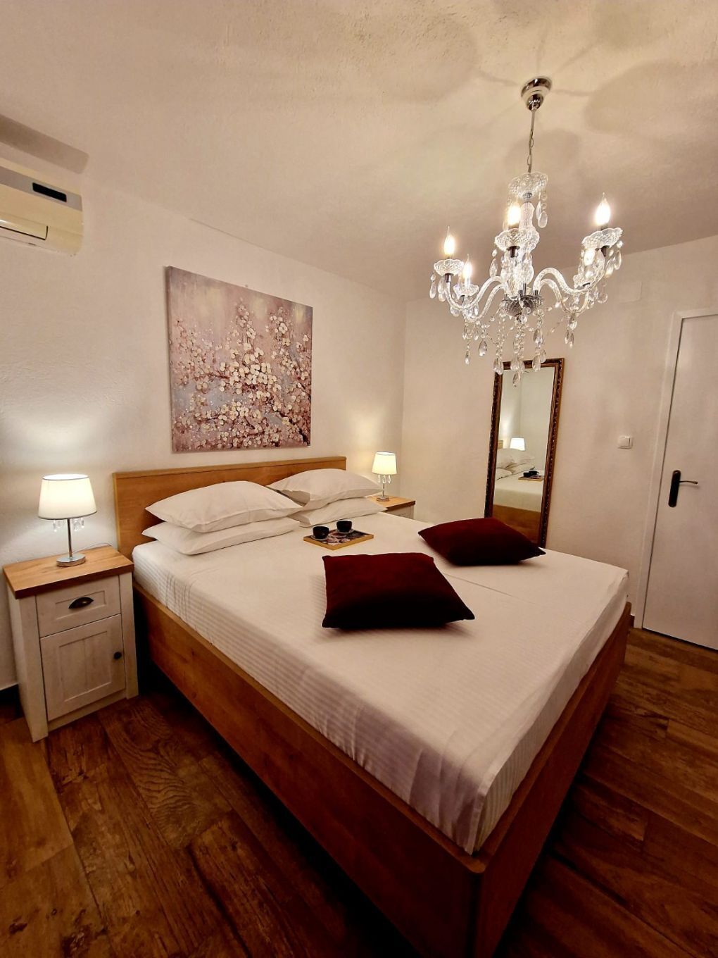 A neatly made bed with white linens, a wooden headboard, and two red pillows. A chandelier hangs above the bed, and a painting adorns the wall.