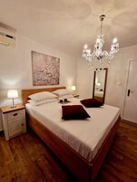 A neatly made bed with white linens, a wooden headboard, and two red pillows. A chandelier hangs above the bed, and a painting adorns the wall.