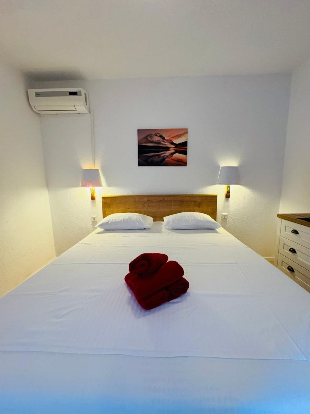 A hotel room with a bed, two bedside lamps, a wall-mounted air conditioner, and a painting above the bed.