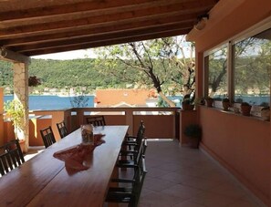 A covered outdoor dining area with a wooden table and chairs, overlooking a scenic view of a body of water and hills. - Apartment and Rooms Mila (Rab)