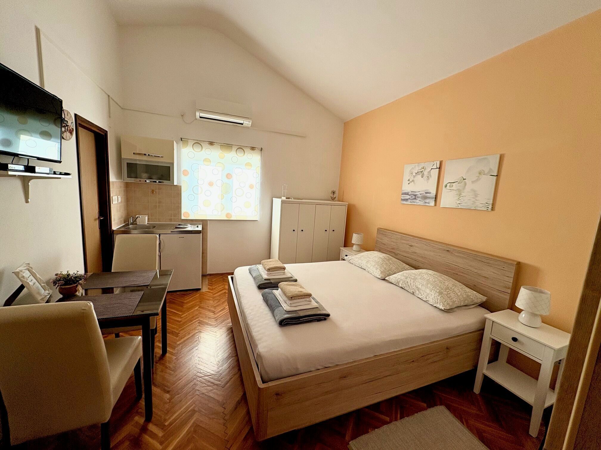 Ein kompaktes Hotelzimmer mit Bett, Nachttisch, kleinem Schreibtisch mit Stuhl, einer Küchenzeile und einem Fernseher.