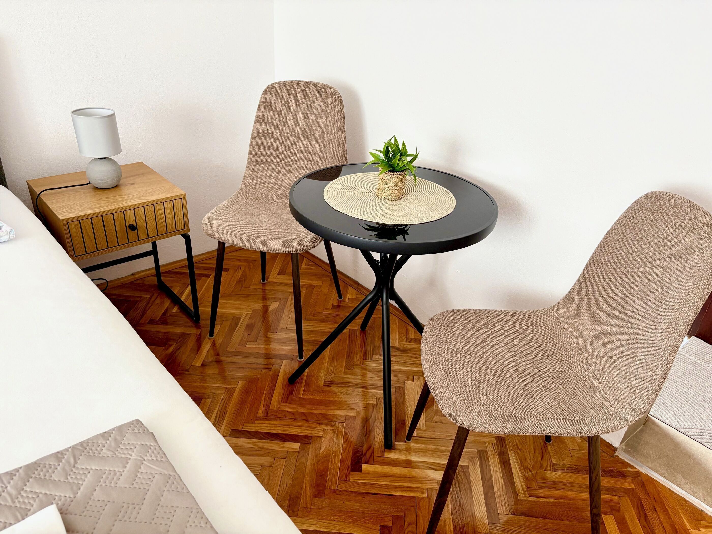 Ein Zimmer mit Holzdielenmuster-Boden, einem runden schwarzen Tisch mit gewebter Tischmatte, zwei beigen Stühlen, einem kleinen Holznachttisch mit Lampe und einer Topfpflanze.