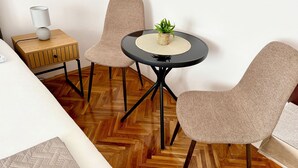A room with a wooden herringbone floor, a round black table with a woven placemat, two beige chairs, a small wooden side table with a lamp, and a potted plant. - Apartments Ognjen (Tisno)
