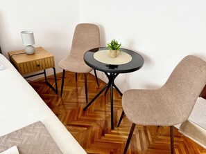 A room with a wooden herringbone floor, a round black table with a woven placemat, two beige chairs, a small wooden side table with a lamp, and a potted plant. - Apartments Ognjen (Tisno)
