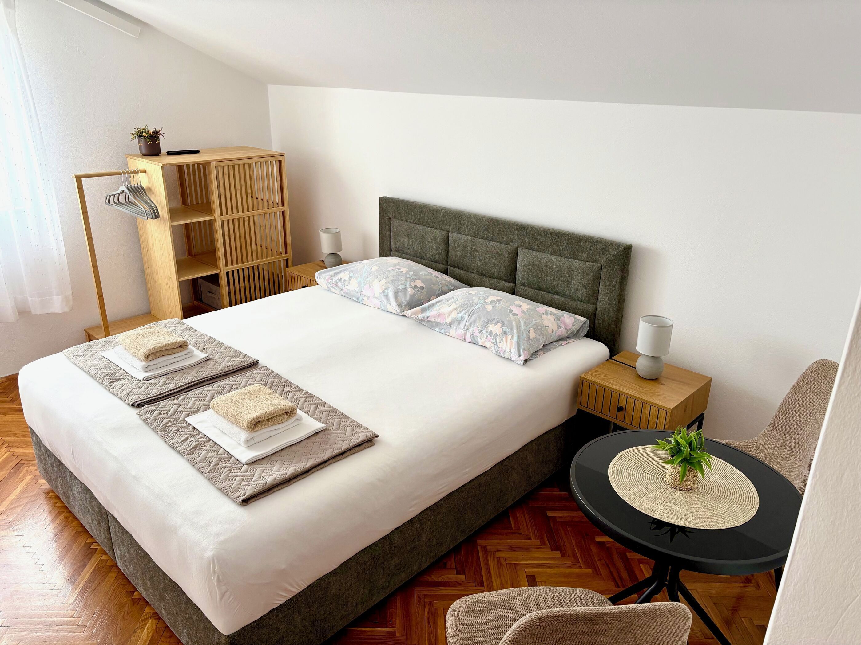 A neatly made bed with pillows, a bedside table with a lamp, a small round table with a plant, and a wooden wardrobe.