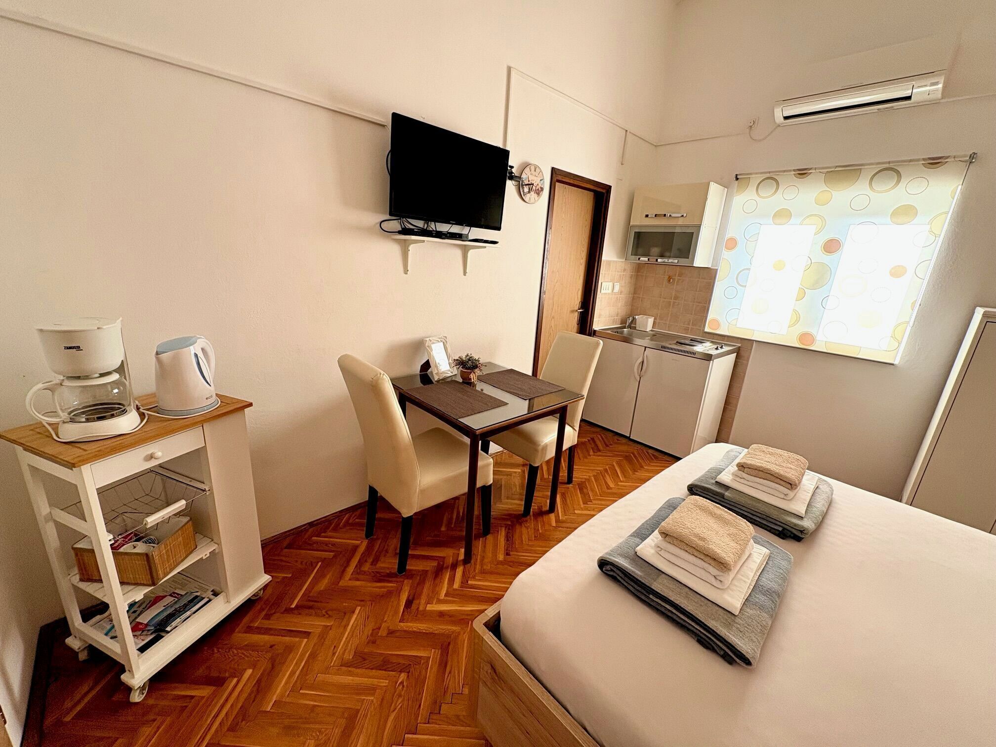 A compact hotel room with a bed, a desk with a chair, a kitchenette, and a television mounted on the wall.