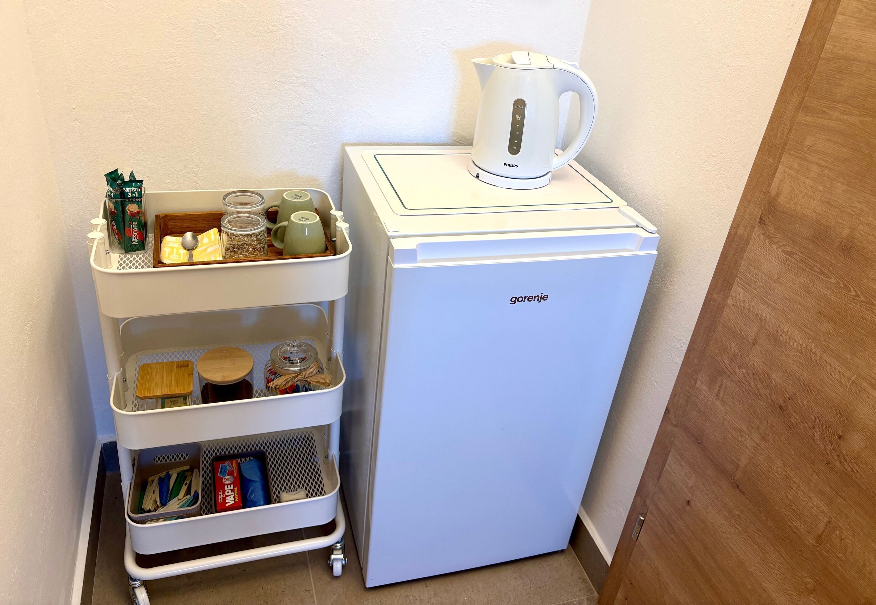 A white refrigerator with a kettle on top, a white cart with storage containers, and a wall-mounted dispenser.
