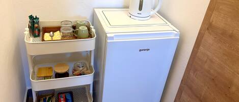 A white refrigerator with a kettle on top, a white cart with storage containers, and a wall-mounted dispenser.