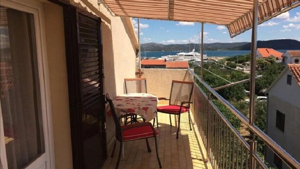A balcony with a table and chairs, overlooking a body of water and buildings. - Apartments Ognjen (Tisno)