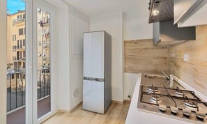 Una cocina compacta con estufa a gas integrada, un refrigerador blanco y una puerta corrediza de vidrio que da a un balcón con vistas a edificios cercanos.