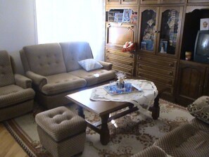 Ein Wohnzimmer mit Sofa, Sessel, Hocker, Couchtisch und einem Schrank mit diversen Gegenständen.