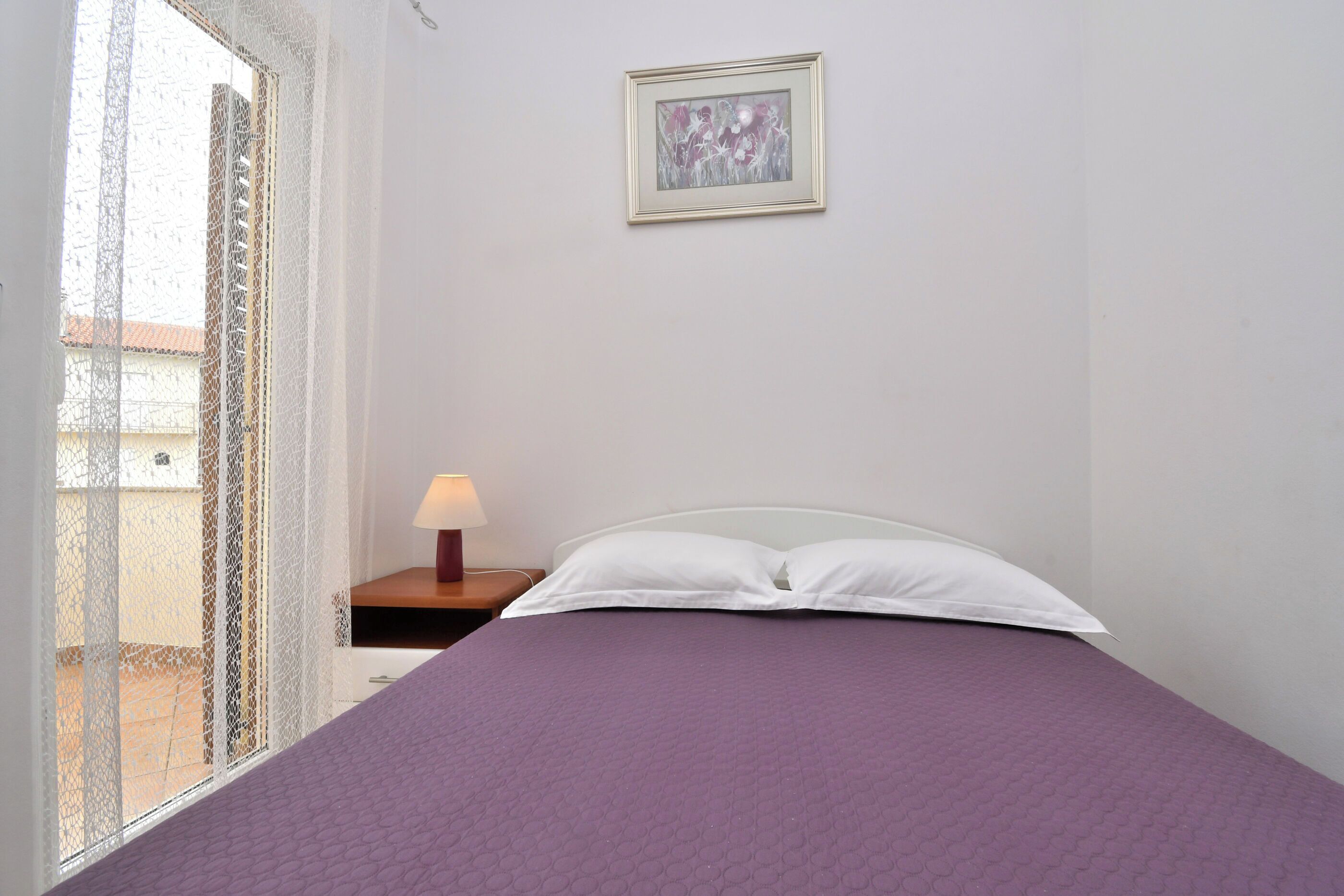 A bedroom with a purple bedspread, a white pillow, a framed picture on the wall, and a window with a patterned curtain.