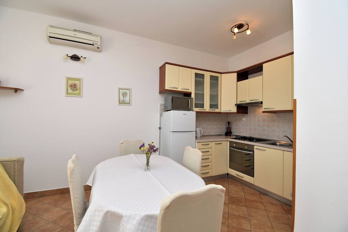 Een kleine keuken met witte kastjes, een witte koelkast en een fornuis. Er is een eettafel met een witte tafelkleed en stoelen.