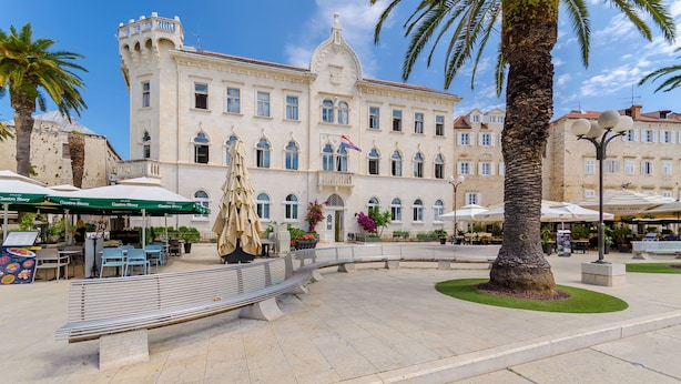 Una piazza pubblica con un grande edificio con finestre ad arco e una fontana centrale.