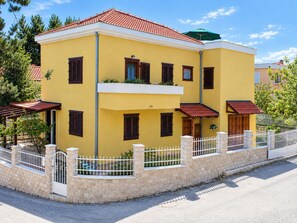 A two-story building with a yellow facade, multiple windows with shutters, a balcony, and a white fence surrounding the property. - Apartment Mana (Split)