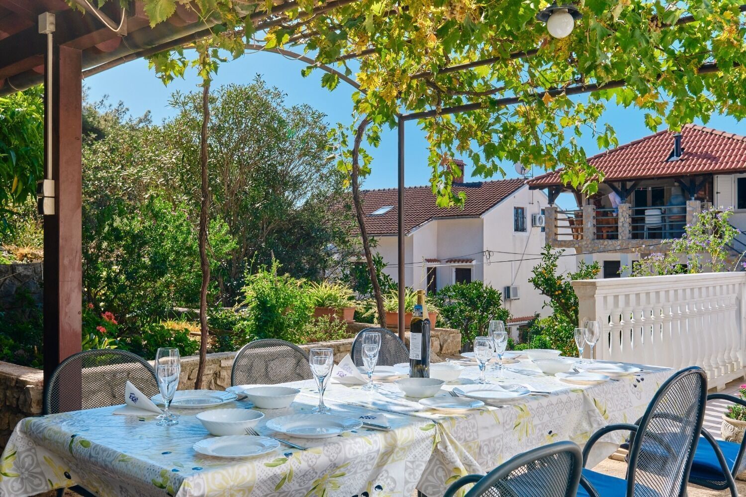 An outdoor dining area with a table set for two, surrounded by greenery and a building in the background.