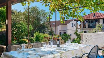 An outdoor dining area with a table set for two, surrounded by greenery and a building in the background.