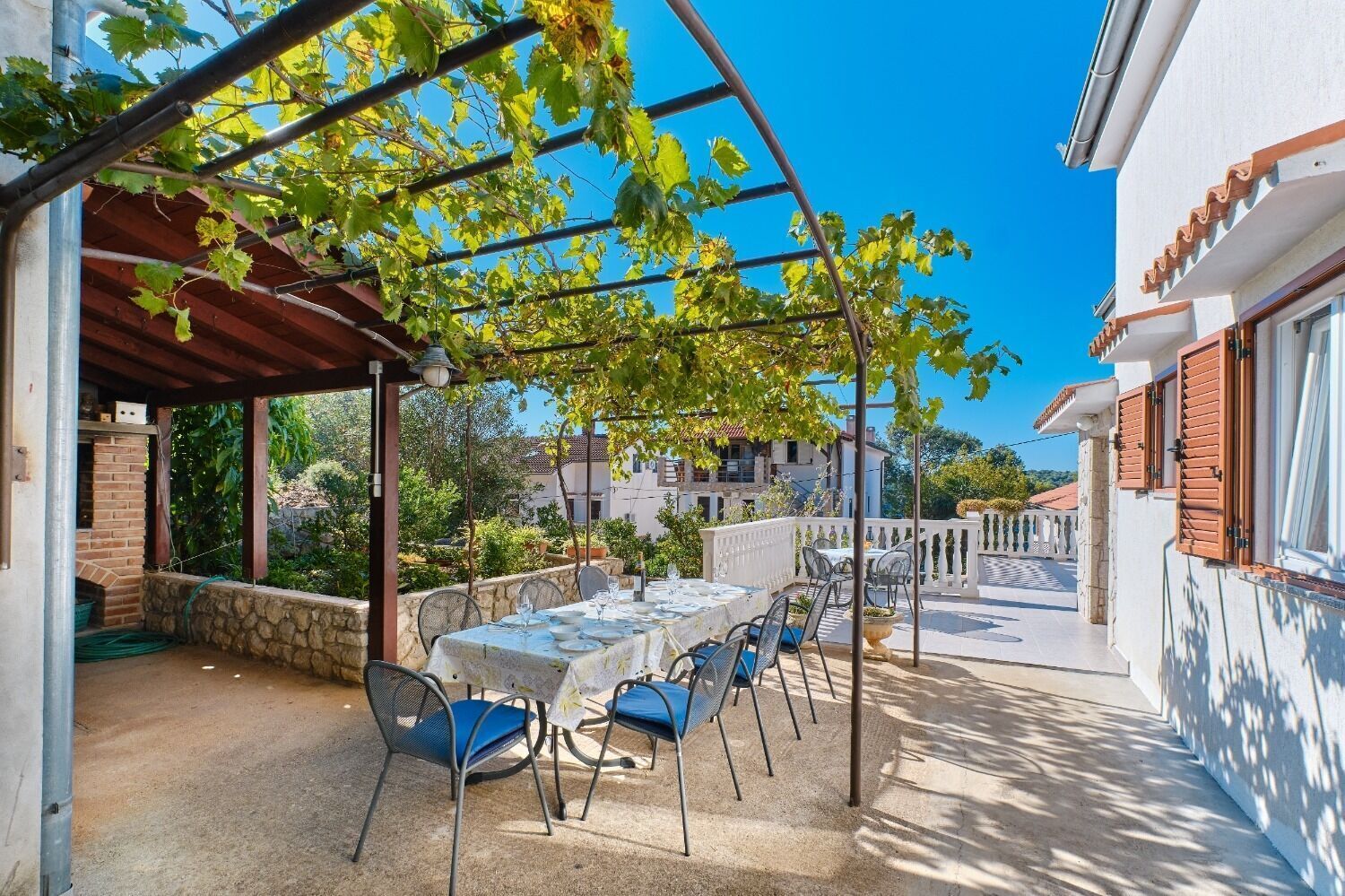 Een overdekt buitenrestaurant met een tafel voor vier personen, omringd door stoelen, onder een pergola met wijnranken.