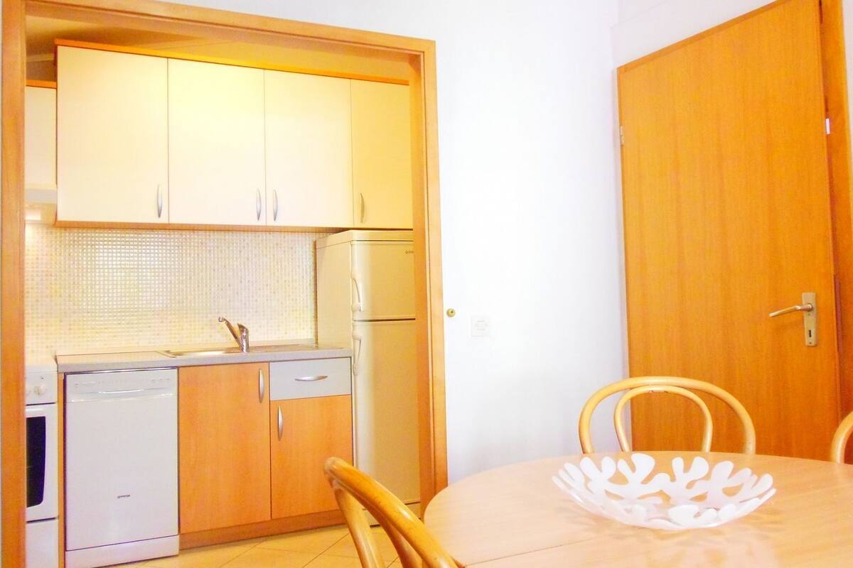 A compact kitchen with wooden cabinets, a refrigerator, and a dishwasher. There is a dining table with a white paperweight on it.