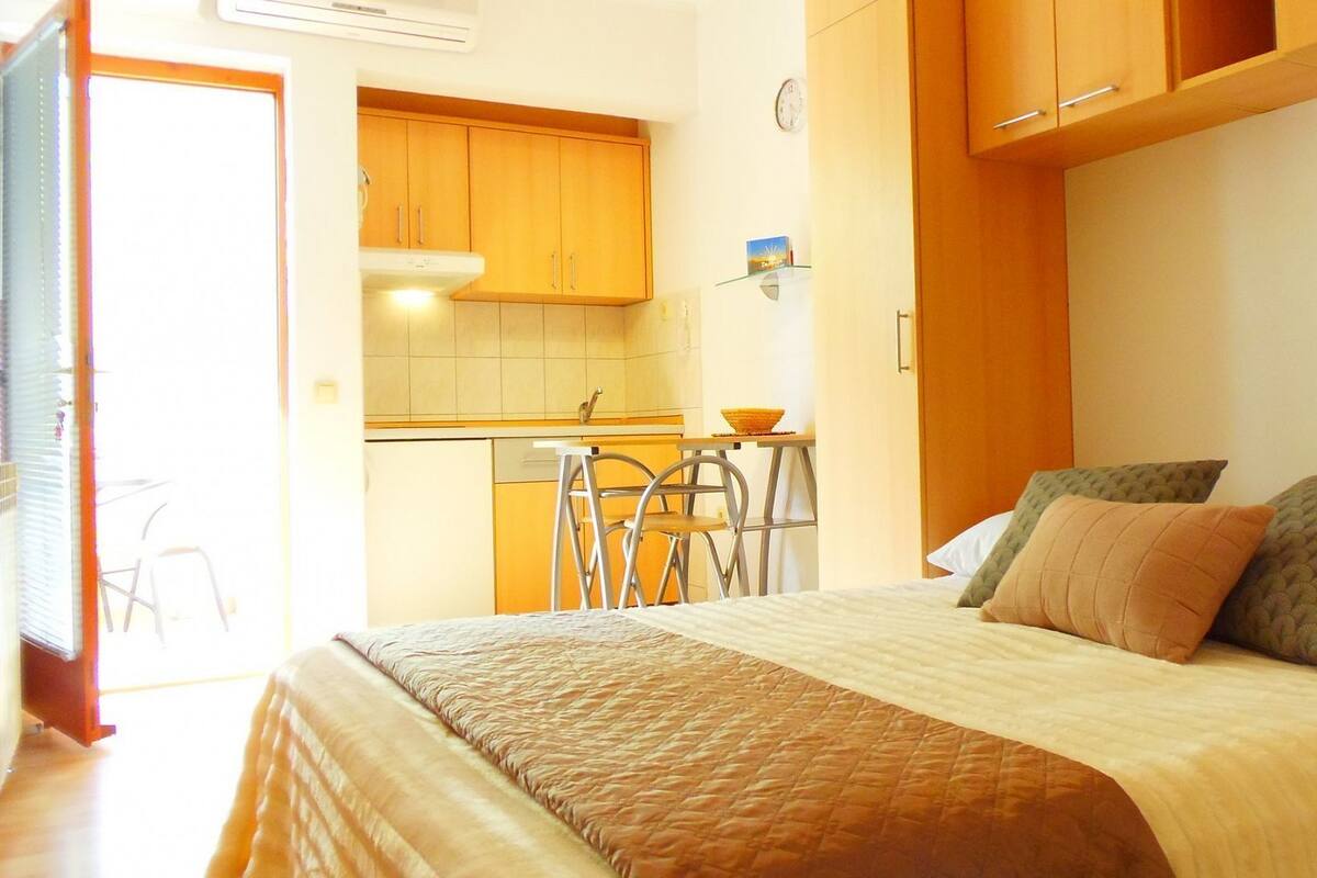 A bedroom with a bed, pillows, and a small kitchenette in the background.