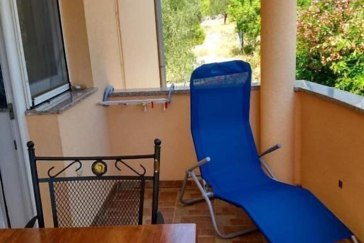 A balcony with a wooden table and a blue lounge chair, overlooking a hilly landscape with greenery.