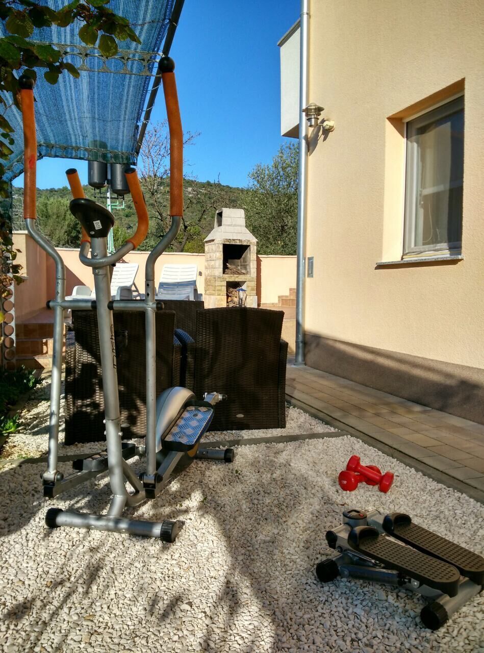 An outdoor exercise area with a stationary bike, a rowing machine, and dumbbells on a gravel surface.