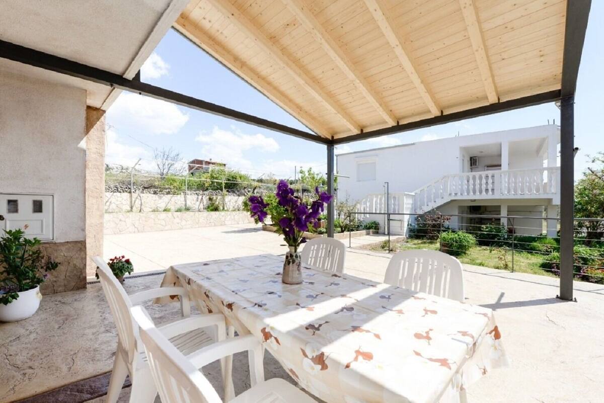 A covered outdoor dining area with a table set for two, surrounded by white chairs, a vase of purple flowers, and a view of a white building with a balcony.