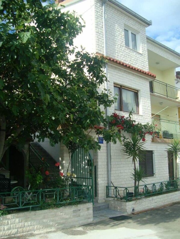 A two-story building with a white brick facade, a balcony on the upper floor, and a green metal gate at the entrance. - Apartment Daria Jure (Trogir)