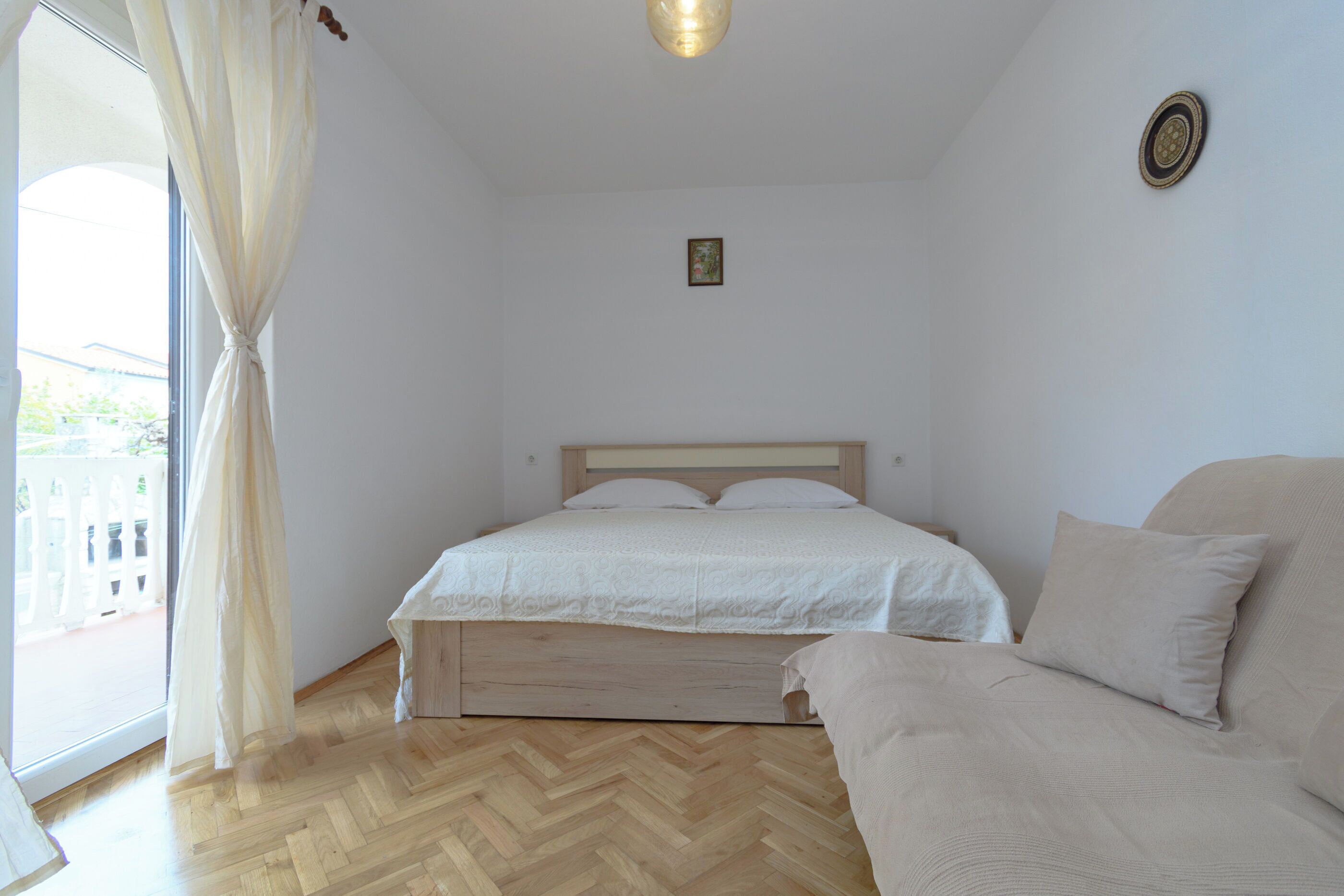 Une chambre à coucher avec un lit, un canapé, une fenêtre avec des rideaux, un luminaire et un petit tableau accroché au mur.