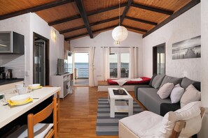 A spacious living area with a wooden ceiling, a dining table with chairs, a sofa, a TV, and a view of the sea through large windows. - Apartment Vlatkica (Jasenice)