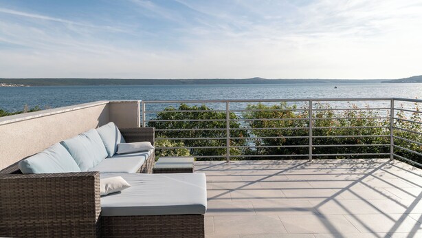 A balcony with a wicker sofa and cushions, overlooking a body of water and greenery. - Apartment Vlatkica (Jasenice)