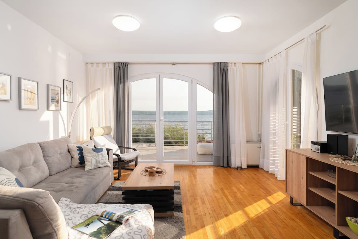 A living room with a sofa, coffee table, TV, and a view of the sea through large windows.