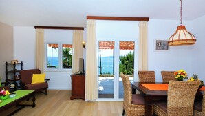 A room with a wooden floor, a sofa, a dining table with wicker chairs, a view of the sea through a glass door, and a hanging light fixture. - House Sreca (Okrug)