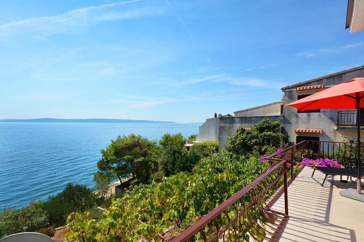 Une propriété située sur la côte, dotée d’un balcon donnant sur la mer, d’un parasol rouge et d’un jardin agrémenté de fleurs violettes.