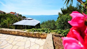 A stone patio with a pergola, lush greenery, and a view of the sea. - House Sreca (Okrug)