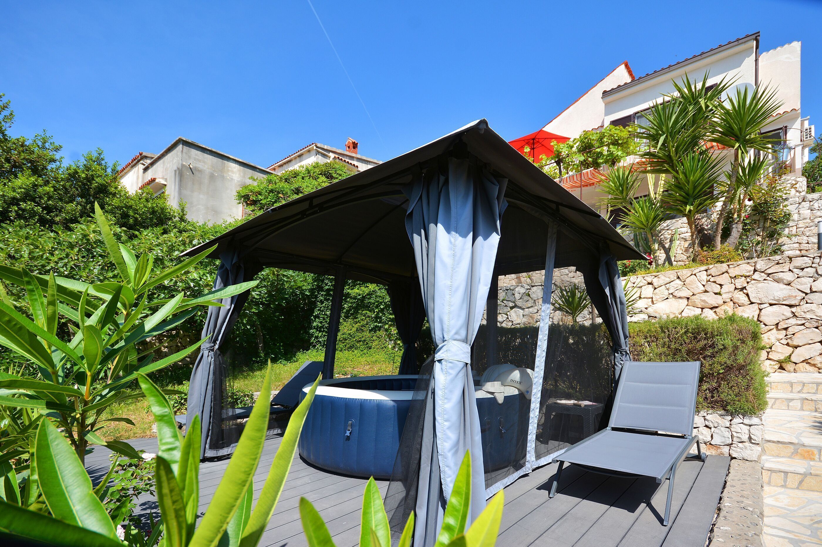 A covered outdoor seating area with a hot tub, surrounded by greenery and a stone wall.