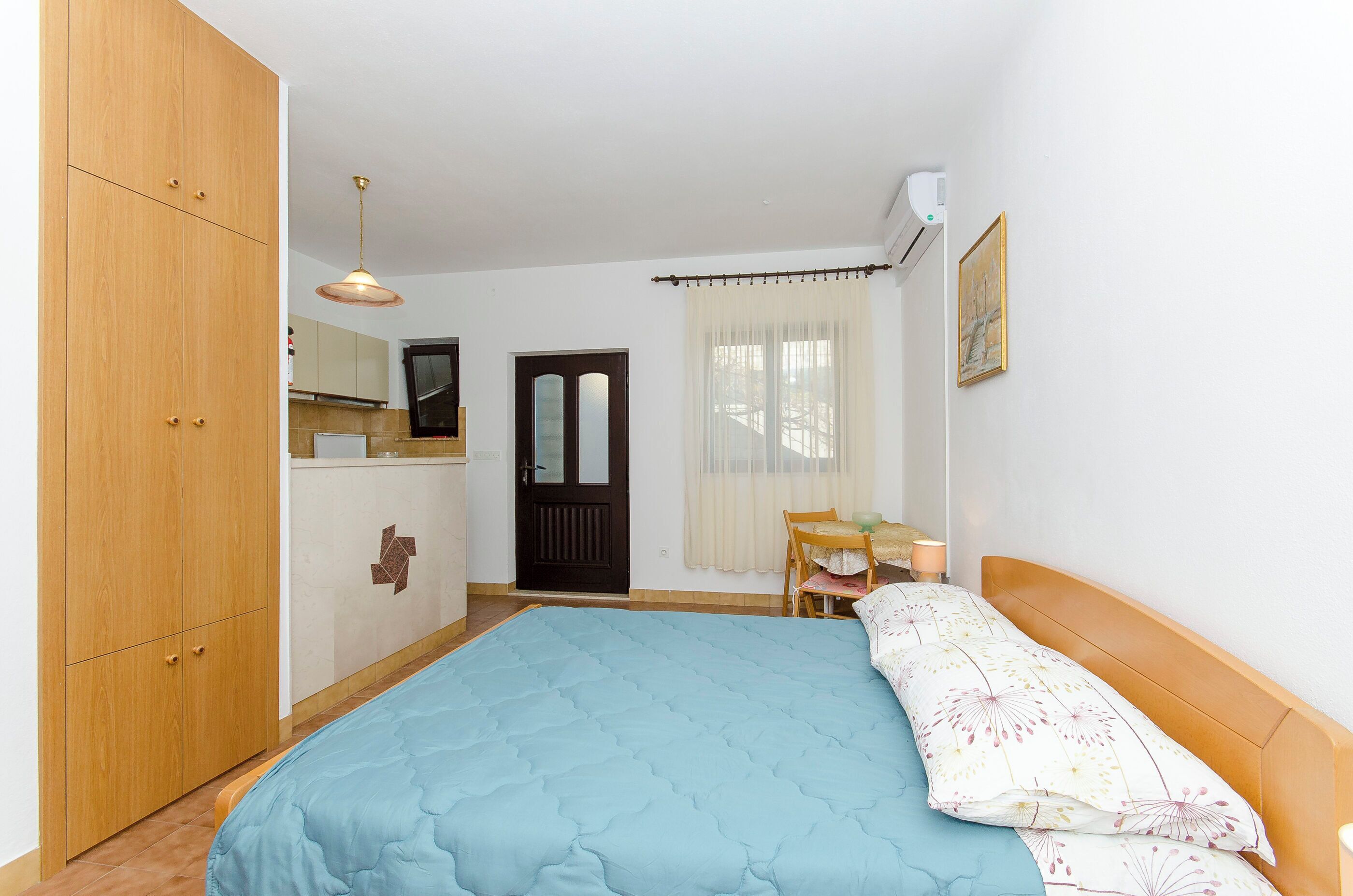A bedroom with a bed, wooden wardrobe, a small table with a chair, a window with curtains, and a wall-mounted air conditioner.