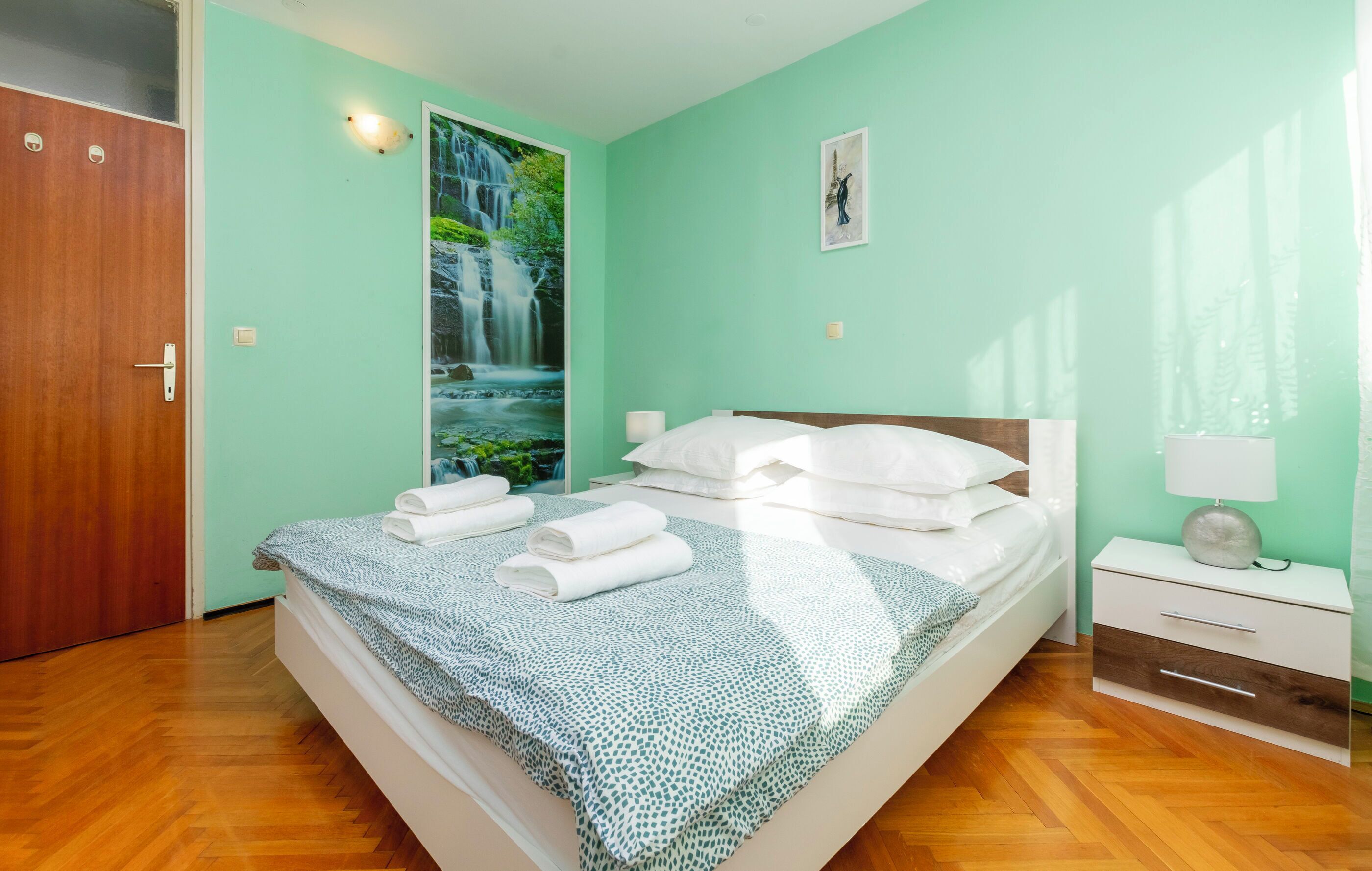 A bedroom with a bed, a nightstand, a wall-mounted lamp, a door, and a large window with a waterfall view.