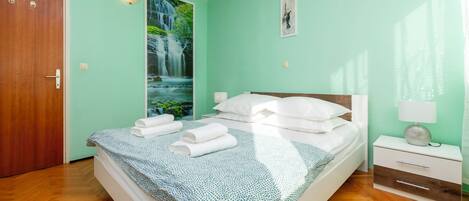 A bedroom with a bed, a nightstand, a wall-mounted lamp, a door, and a large window with a waterfall view.