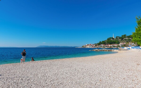 A pebble beach with clear blue water, a small town on a hill, and a lighthouse in the distance. - Apartment Livr (Podgora)