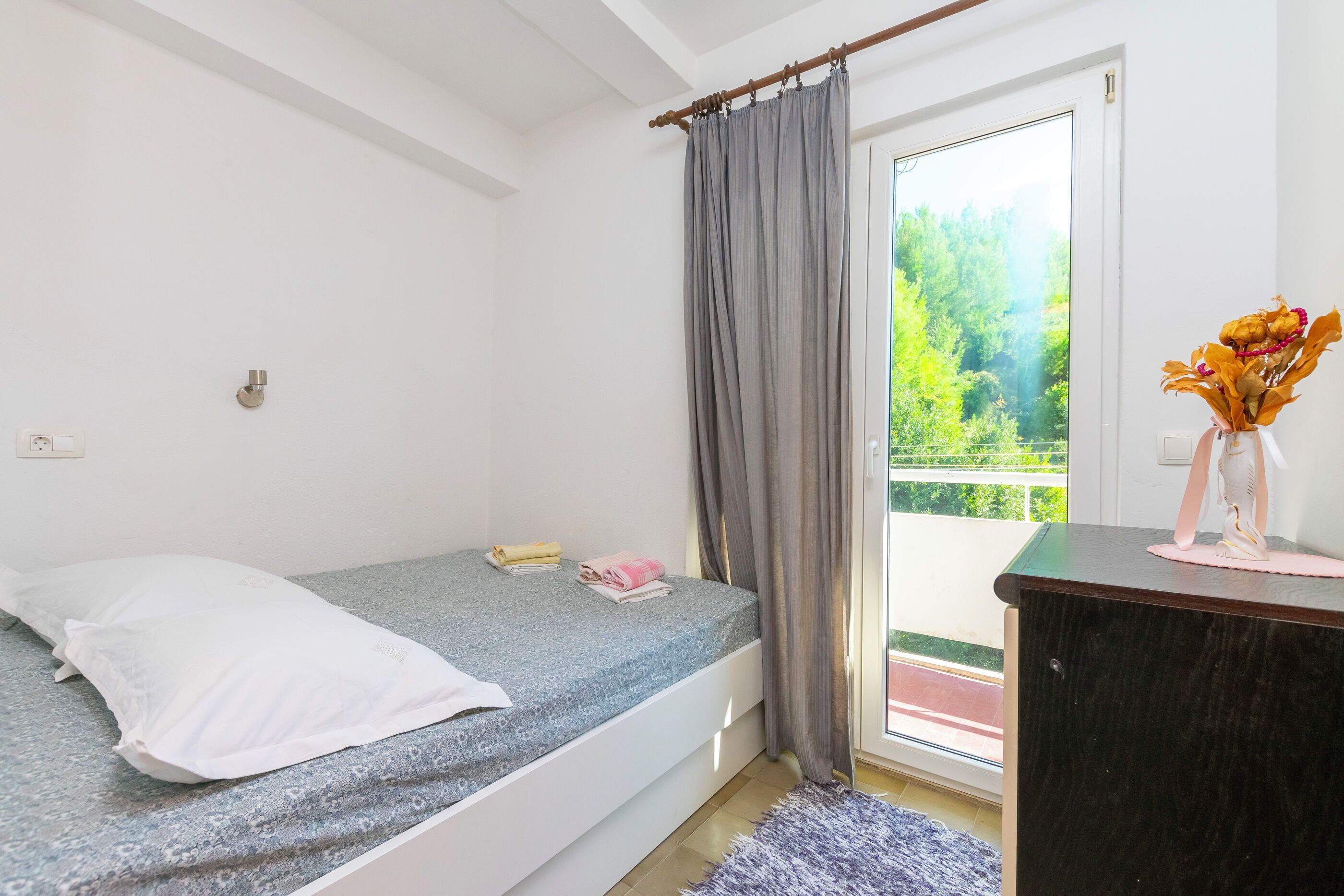 Une petite chambre avec un lit simple, un matelas gris, un oreiller blanc et une petite table de chevet sur laquelle est posé un vase contenant des fleurs séchées.