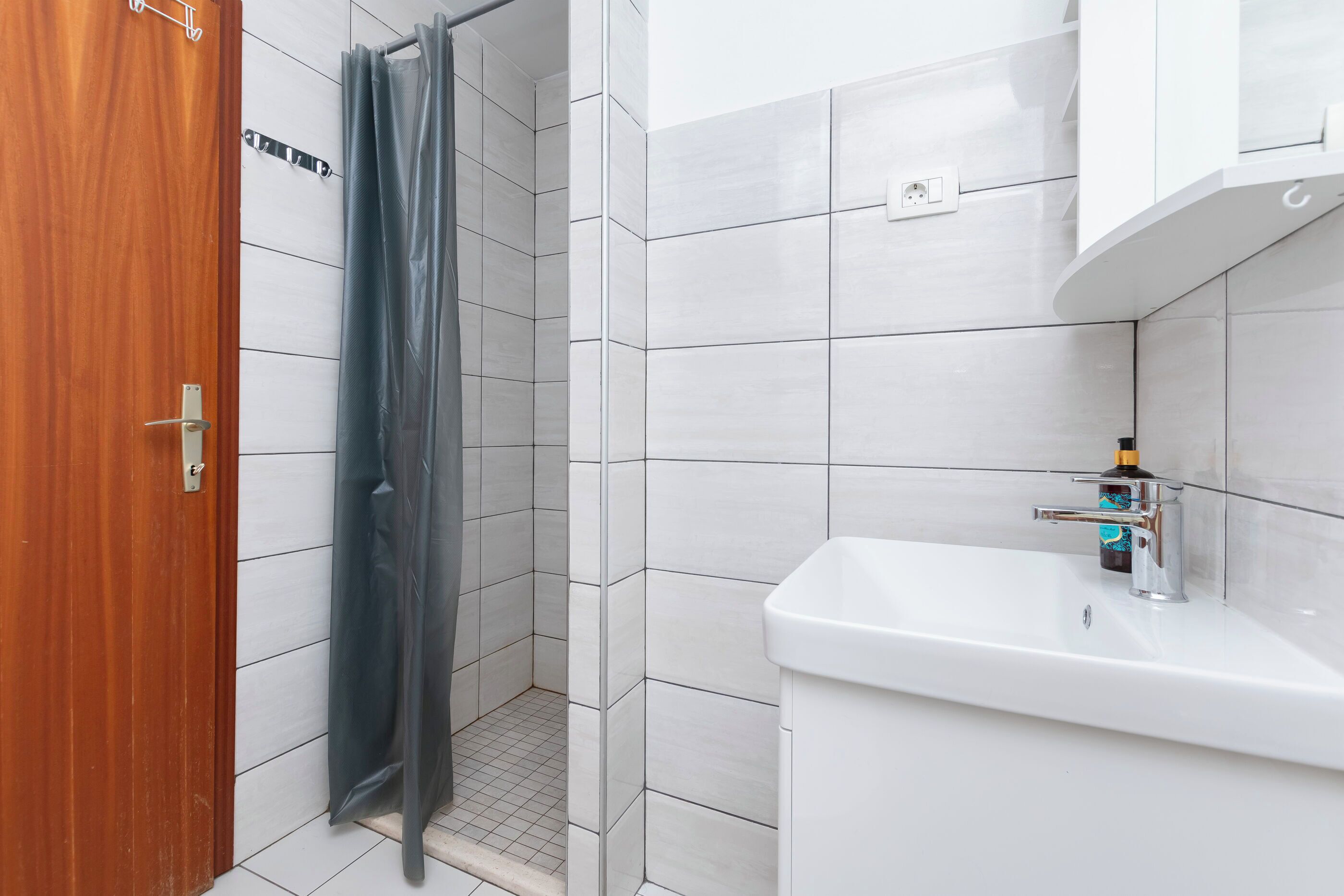 Een badkamer met een houten deur, een douche met een grijs gordijn, witte betegelde muren en een witte wastafel met een kraan.