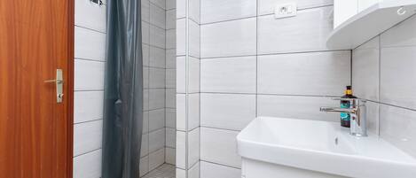 Een badkamer met een houten deur, een douche met een grijs gordijn, witte betegelde muren en een witte wastafel met een kraan.