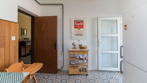 Een compacte keuken met een houten kastje, een eettafel met geruit tafelkleed, een wandplank met keukenspullen, en een koelkast.