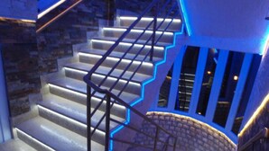 Eine Treppe mit beleuchteten Handläufen und Stufen, die zu einem Zwischenpodest mit einer Steinmauer und Fenstern führt.